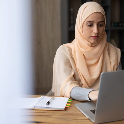 focused muslim woman in hijab sitting at table with laptop and organizer